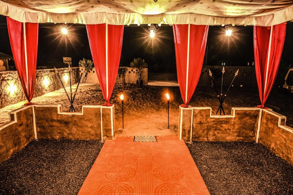 Explore Overnight Desert Camp Jaisalmer entrance at night — red velvet drapes, hanging lanterns and fire torches on sand